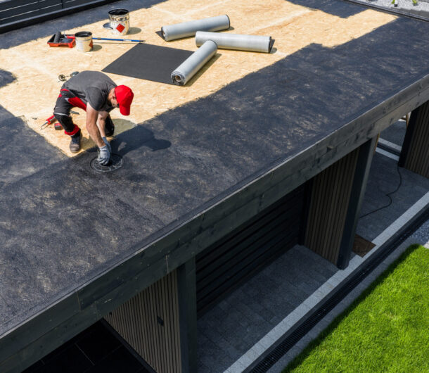 A construction worker is installing EPDM roofing material on a contemporary home under clear skies, surrounded by tools and prepared materials for the roofing project.