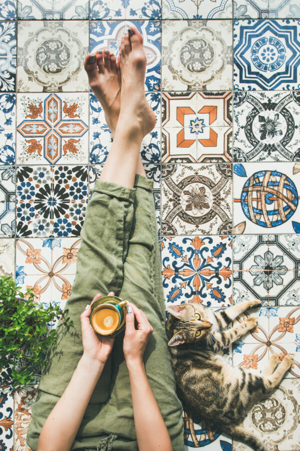 Lazy morning on terrace. Flat-lay of womans legs in cosy linen pants, plant, cat and cup of coffee in hand over colorful moroccan tile floor, top view